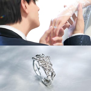 Puede incluir: Un anillo plateado con diseño floral, con múltiples gemas transparentes. El anillo se muestra en un estudio, con manos en el fondo, una de ellas con un anillo similar. El anillo es una pieza de joyería.