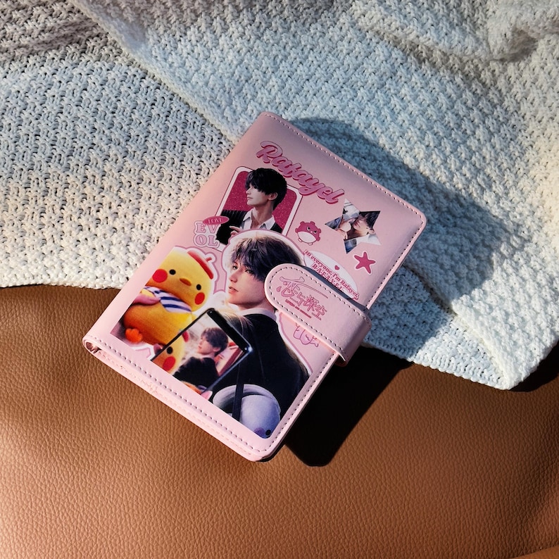 May include: Pink wallet with a flap closure. The wallet features images of a person, a cartoon chick, and text. The word "Patayel" is written in cursive on the top of the wallet. The wallet is on a brown surface.
