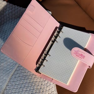 May include: A pink planner with a silver binder and a light blue grid-lined notepad. The planner is open, revealing card slots and a pen holder. The notepad is secured by a pink strap with text. The planner is on a brown surface.