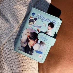 May include: A light blue wallet with a magnetic closure. The wallet features illustrations of anime characters and the name "Zayne". The wallet has a stitched border and a strap closure.