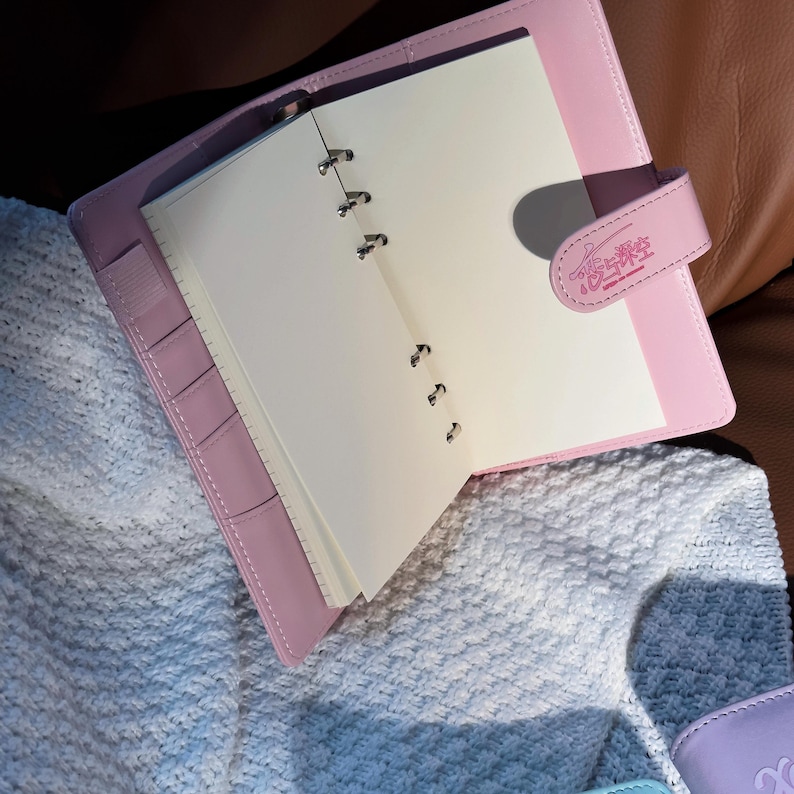May include: A light pink leather-bound planner with a snap closure. The planner is open, revealing blank white pages. The cover features the text "恋与深空" in pink. The planner has a pen holder and several pockets.