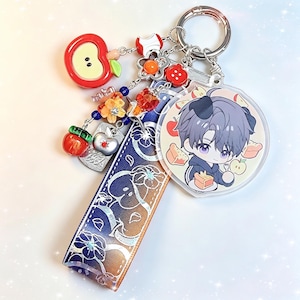 May include: A silver keychain with multiple charms. Charms include a red apple, a flower, and a circular charm with an anime character. A blue and white patterned ribbon is also attached.