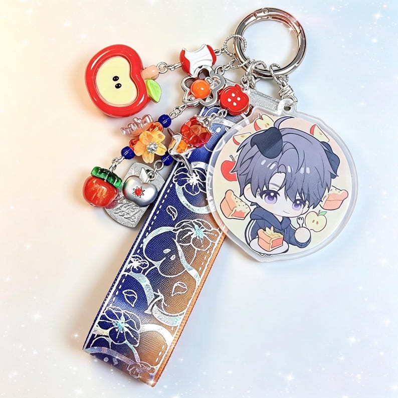 May include: A silver keychain with multiple charms. Charms include a red apple, a flower, and a circular charm with an anime character. A blue and white patterned ribbon is also attached.