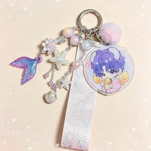 May include: A decorative keychain featuring a cartoon character with purple hair and a pink outfit, holding two yellow chicks. The keychain includes a pink pom-pom, a mermaid tail charm, and various sea-themed charms, such as starfish and pearls.
