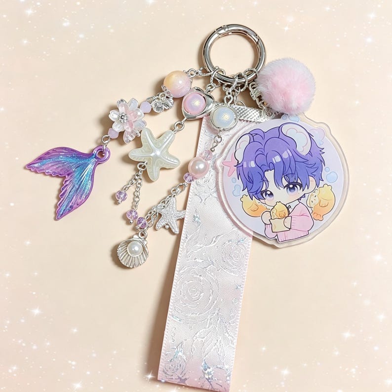 May include: A decorative keychain featuring a cartoon character with purple hair and a pink outfit, holding two yellow chicks. The keychain includes a pink pom-pom, a mermaid tail charm, and various sea-themed charms, such as starfish and pearls.
