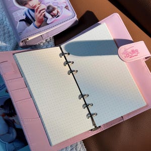 May include: A pink binder notebook with a dot grid paper insert. The notebook features a snap closure and a small image on the cover. A smaller, matching notebook is visible in the background.