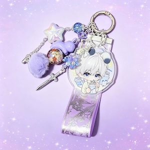 May include: A decorative keychain with a silver ring, featuring a clear charm with an anime character, a purple ribbon, and various charms. Charms include a star, a bunny, a pom-pom, a sword, and beads in shades of purple and blue.