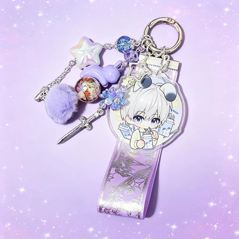 May include: A decorative keychain with a silver ring, featuring a clear charm with an anime character, a purple ribbon, and various charms. Charms include a star, a bunny, a pom-pom, a sword, and beads in shades of purple and blue.