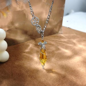 Puede incluir: Un collar steampunk plateado con un colgante amarillo en forma de lágrima. El collar presenta una cadena con detalles en forma de engranaje y un colgante envuelto en alambre. El colgante mide unos 5 cm de largo.