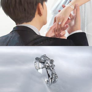 Puede incluir: Un anillo plateado con diseño de estrella y cristales. El anillo presenta un elemento central en forma de estrella, rodeado de cristales más pequeños y detalles decorativos. El anillo se muestra sobre una superficie blanca.