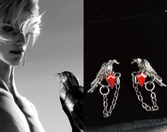 May include: Silver-toned raven earrings with red gemstone accents and dangling chains. The earrings are detailed with a bird perched on a branch, and are set against a black background. The image also features a person and a raven.