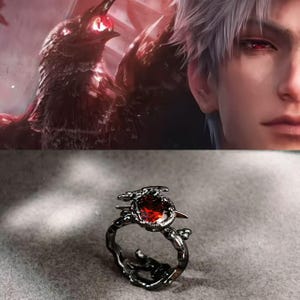 Puede incluir: Un anillo negro con una gran gema roja ovalada. El anillo tiene un diseño detallado inspirado en la naturaleza con un motivo de pájaro y rama. El fondo presenta una ilustración digital de un cuervo y una persona con ojos rojos.