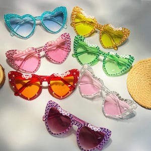 May include: A set of ten heart-shaped sunglasses with rhinestones and different coloured lenses. Each pair has a different word on the frame, including "Lover", "Shake It Off", "Eras Tour", and "Bad Blood".