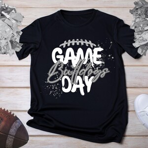 May include: Black t-shirt with the words "GAME Bulldogs DAY" in white and gray, with a football graphic. A brown football and white sneakers are also visible. The shirt is on a light wood surface.