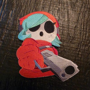 Puede incluir: Un parche bordado que muestra un personaje con una máscara blanca, grandes cuencas oculares negras y cabello color verde azulado. El personaje viste una prenda roja con capucha y sostiene una pistola gris apuntando hacia adelante con hilo denso.