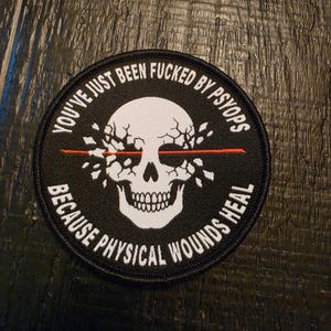 May include: A circular black embroidered patch featuring a white skull with a cracked forehead and a horizontal red line across the eyes. The text reads: YOU'VE JUST BEEN FUCKED BY PSYOPS BECAUSE PHYSICAL WOUNDS HEAL in white capital letters.