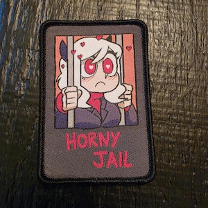 May include: A rectangular embroidered patch with a black border featuring an anime-style character with white hair and heart-shaped eyes behind grey bars. The background is pink with small red hearts. The text HORNY JAIL is embroidered in red.