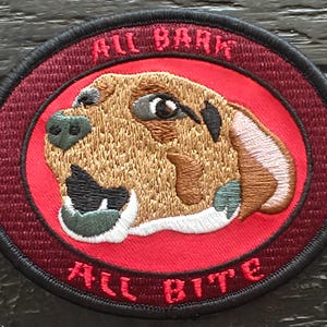 May include: Round embroidered patch with a dog's head design. The text "ALL BARK" and "ALL BITE" are embroidered above and below the dog's head. The dog has brown fur and a black nose, set against a red background.