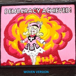 May include: Embroidered patch featuring the text "DEMOCRACY ACHIEVED!" above an anime-style character in a pink dress, set against a yellow and red explosion. The patch is bordered in black, with "WOVEN VERSION" at the bottom.