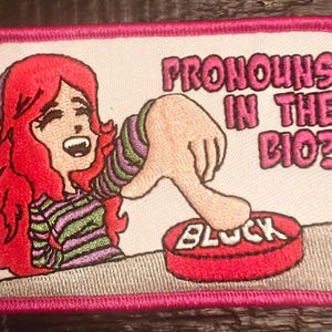 Può includere: Una toppa ricamata rosa e bianca con una donna dei cartoni animati che preme un pulsante rosso che dice "Blocca". Il testo sopra il pulsante dice "Pronomi nella bio?"