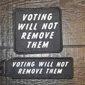 Può includere: Due toppe nere con testo bianco "VOTING WILL NOT REMOVE THEM". Una è quadrata, l'altra rettangolare con una clip nera. Sono su una superficie di legno scuro.
