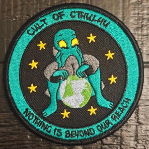 Puede incluir: Parche redondo con borde turquesa y texto "CULT OF CTHULHU" y "NOTHING IS BEYOND OUR REACH". Presenta una criatura turquesa sosteniendo un globo terráqueo, rodeada de estrellas amarillas, sobre un fondo negro.