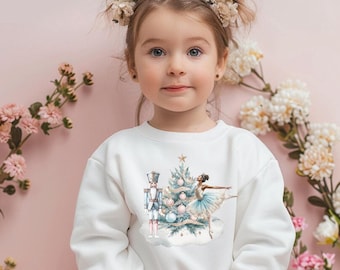 May include: White sweatshirt featuring a watercolor design of a Christmas tree, a nutcracker, and a ballerina. The design includes light blue and pink accents. The sweatshirt is worn by a young child with floral hair accessories.