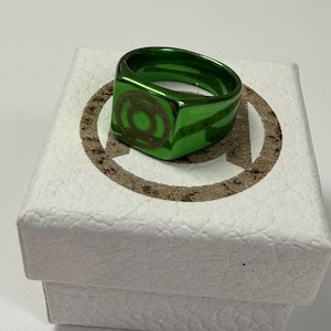 May include: A metallic green ring featuring a circular Green Lantern emblem engraved on the flat top surface. The ring has a polished, reflective finish and is displayed resting on a white textured box with a circular logo printed on the lid.