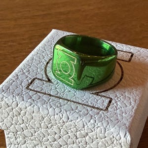 May include: A metallic green ring featuring the circular Green Lantern logo engraved on the front face. The ring is displayed on top of a white, textured square box that also features the same circular Green Lantern logo embossed on its top surface.