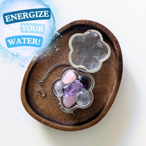 May include: A stainless steel flower-shaped tea infuser with a chain, filled with pink and purple crystals. The infuser is sitting on a wooden tray. The text "Energize Your Water!" is in a blue circle with water droplets.