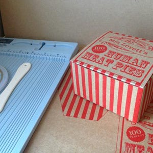 Può includere: Una scatola a strisce rosse e bianche con il testo "Mrs. Lovett's 100 Percent Human Meat Pies" stampato sulla parte superiore. La scatola è appoggiata su una superficie marrone.