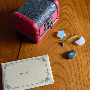 May include: A small wooden box with a floral design and a red lid. The box is open and there are several small objects inside, including a star, a feather, a crescent moon, and a black stone with a gold symbol. The box is sitting on a wooden table with a note that says "Open Last".