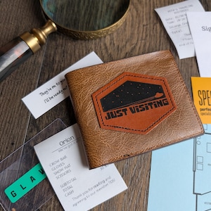 Pode incluir: Uma carteira castanha de duas dobras com um emblema hexagonal de fundo preto, um pequeno gráfico de um OVNI laranja e o texto "JUST VISITING" em letras pretas a negrito. A carteira está rodeada por vários papéis, uma lupa e cartões.