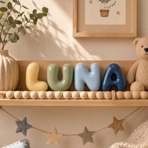 Peut inclure: Quatre lettres en céramique formant le nom LUNA, chacune avec une forme arrondie, lisse et brillante rappelant un ballon. La lettre L est jaune pâle, U est vert sauge, N est bleu clair et A est bleu marine, posées sur une étagère en bois.