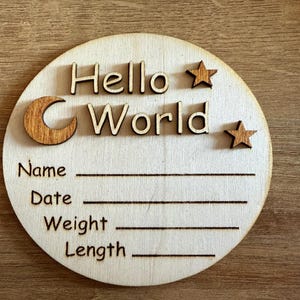May include: A round wooden plaque with the text "Hello World" and two stars and a crescent moon. The plaque has blank lines for a name, date, weight, and length.
