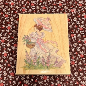 May include: A wooden stamp featuring a girl in a pink dress and a white parasol walking through a field of pink flowers. She is carrying a basket of flowers.
