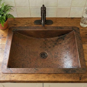 May include: A rectangular copper sink with a hammered texture set into a wooden countertop. A dark bronze tap and a clear glass soap dispenser are visible. A small potted fern sits to the left, and the background is a tiled wall.