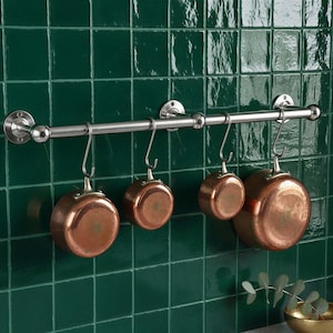 May include: A stainless steel pot rack with four copper pots hanging from it. The rack is mounted on a dark green tiled wall. A small gold bowl with greenery sits on the counter below.