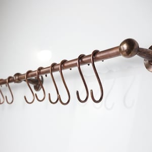 Puede incluir: Una barra de cocina de color cobre con múltiples ganchos en forma de S. La barra está montada en una pared blanca y presenta una bola decorativa en un extremo. Los ganchos están espaciados para colgar utensilios de cocina.