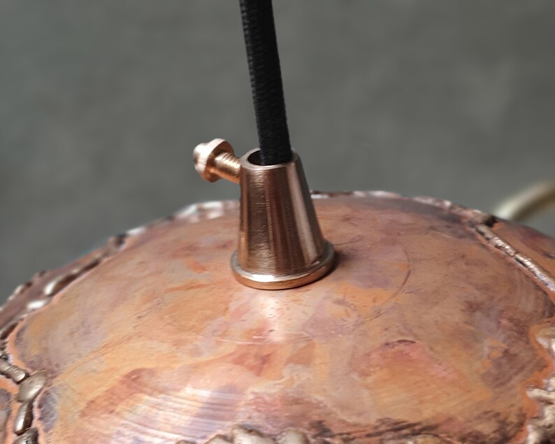 May include: Close-up of a copper pendant light fixture with a brass ceiling mount and black electrical cord.