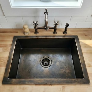 Op de afbeelding: Een rechthoekige, donkerbronzen gootsteen met bijpassende kraan en sproeier. De kraan heeft kruisvormige handgrepen. Een houten schrobborstel staat links van de kraan. De gootsteen is ingebouwd in een licht houten aanrecht.