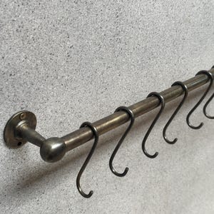 Op de afbeelding: Een messing keukenutensilienrek met een afgerond uiteinde en meerdere haken. Het rek is met twee beugels aan een muur bevestigd.