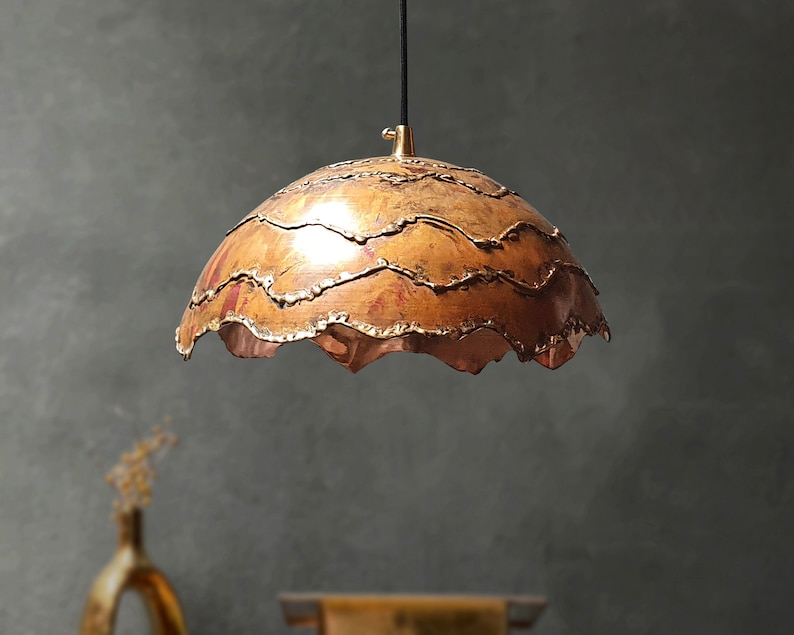 May include: A copper pendant light fixture with a wavy, textured shade. The shade is a warm copper color with hints of red and gold.