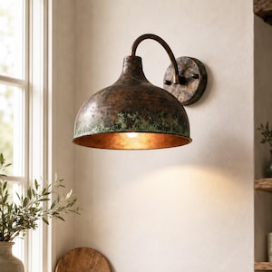 May include: A wall-mounted lamp featuring a dome-shaped metal shade with a weathered, dark brown and oxidized green patina finish. The fixture includes a curved arm connecting the shade to a circular wall plate, emitting a warm light from within.