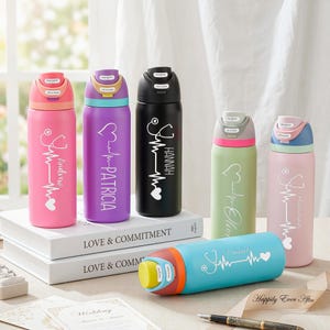 Op de afbeelding: Een verzameling gepersonaliseerde waterflessen in verschillende kleuren, waaronder roze, paars, zwart, groen en blauw. Elke fles heeft een stethoscoopontwerp, een hartslaglijn en een naam. De flessen staan op een witte ondergrond.
