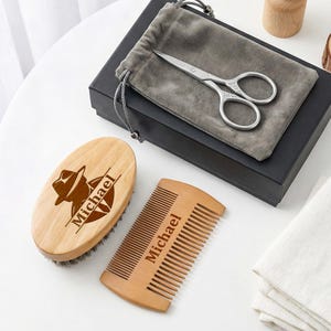 May include: A grooming kit with a wooden beard brush and comb, both engraved with "Michael". A pair of silver scissors and a gray velvet pouch are included. The kit is presented on a white surface with a black box.
