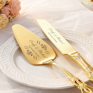 May include: A gold-colored cake server and knife set with ornate handles, resting on a white plate. The server and knife are engraved with the names "Emily & Michael" and the date "September 08, 2025".
