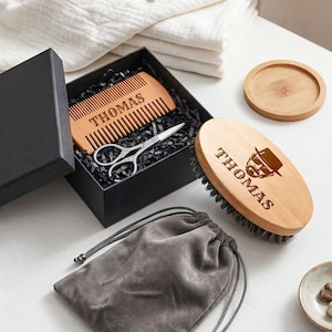 May include: A wooden beard grooming kit with the name "THOMAS" engraved on the comb and brush. The kit includes a comb, scissors, a brush, a drawstring bag, and a small wooden coaster. The brush features a graphic of a man with a hat.