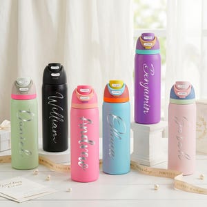 Puede incluir: Una colección de botellas de agua personalizadas y coloridas. Cada botella es de un color diferente, incluyendo verde, negro, rosa, azul, morado y rosa. Los nombres Daniel, William, Andrea, Olivia, Benjamin y Sarah están escritos en las botellas.