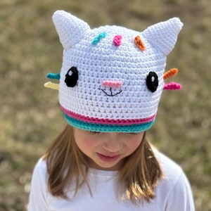 May include: A white crocheted cat hat with colorful accents. The hat has black eyes, a pink nose, and a white smile. The hat is worn by a child with long blonde hair.